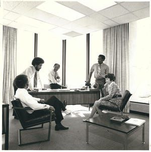 People in a small office meeting around a table, with large windows and curtains in the background.