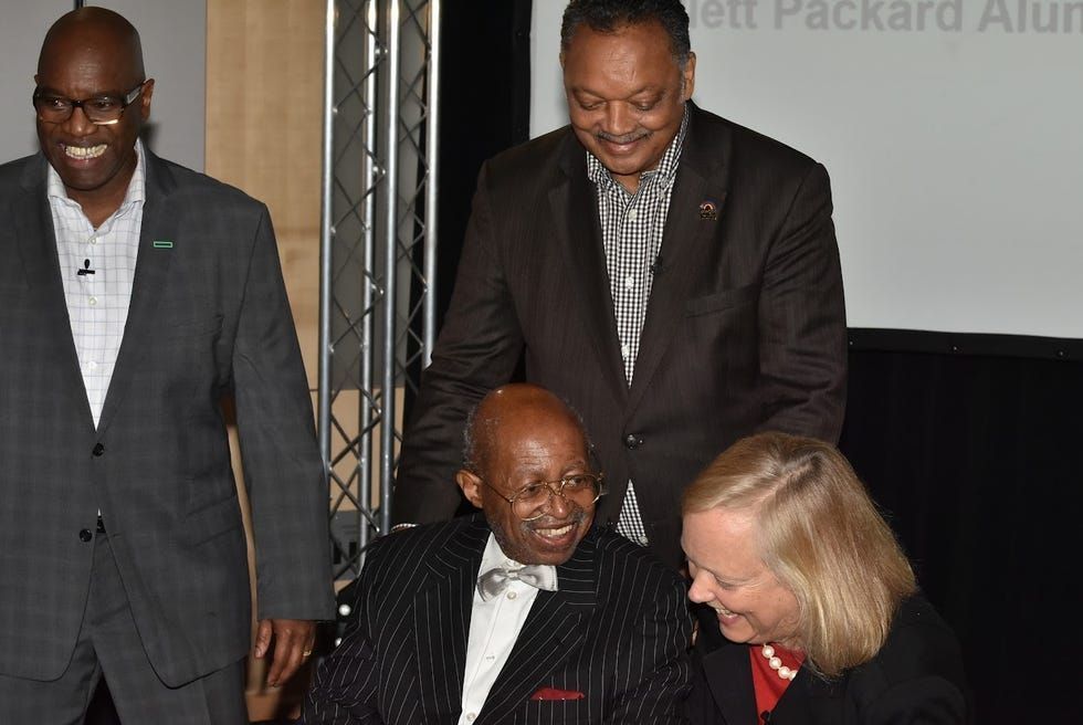 Four people in suits converse onstage at an event, with one seated and smiling.