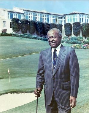 Man in a pinstripe suit standing on a golf course with a large white building in the background