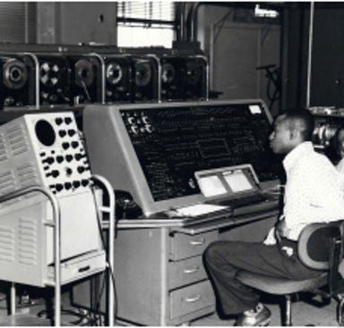 Man seated at a vintage control console with dials and gauges in an industrial equipment room.