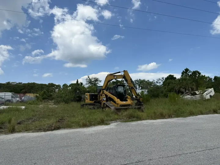 Yellow excavator parked in a grassy field beside a road under a blue, cloudy sky