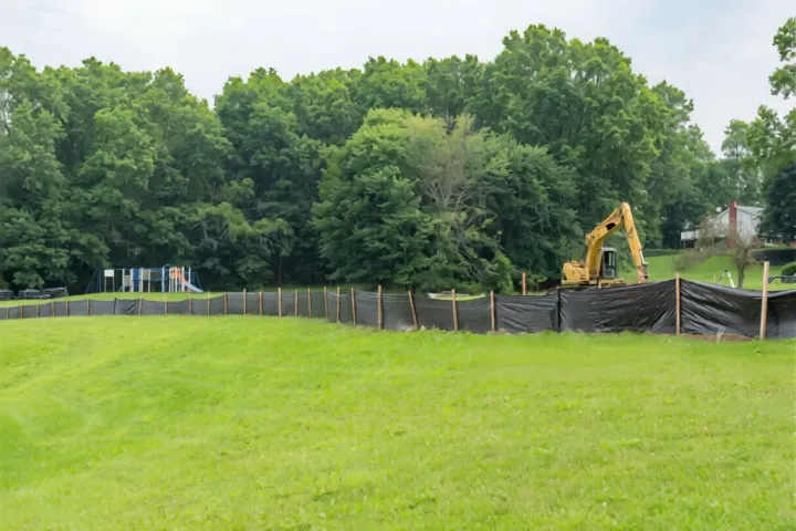 Grassy field beside a tree line with a yellow excavator and black silt fencing under cloudy skies