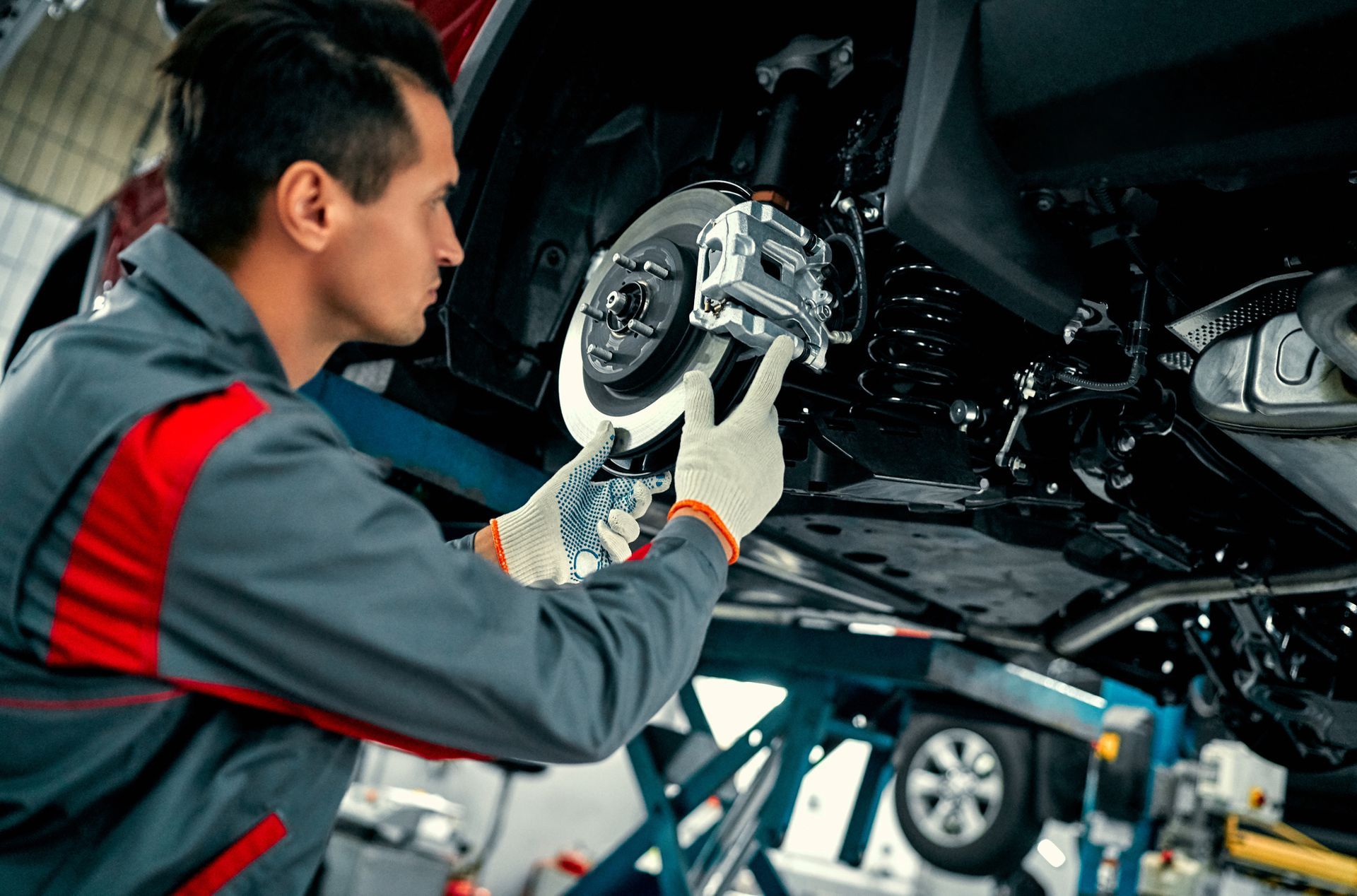 Mechanic in a gray uniform working on a car's brake system, using gloves in a garage.