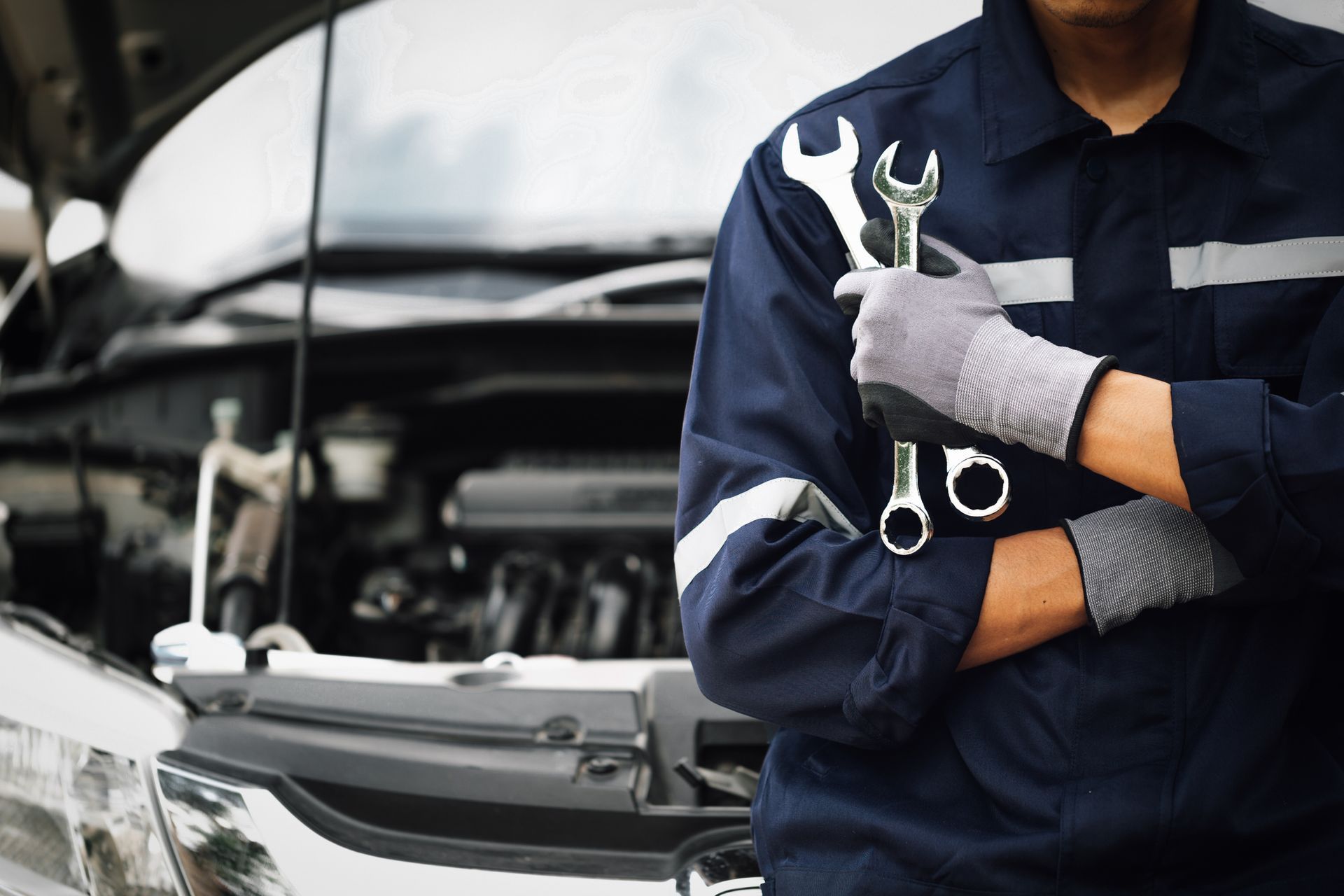Mechanic in blue uniform holding wrenches, standing near a car with open hood.