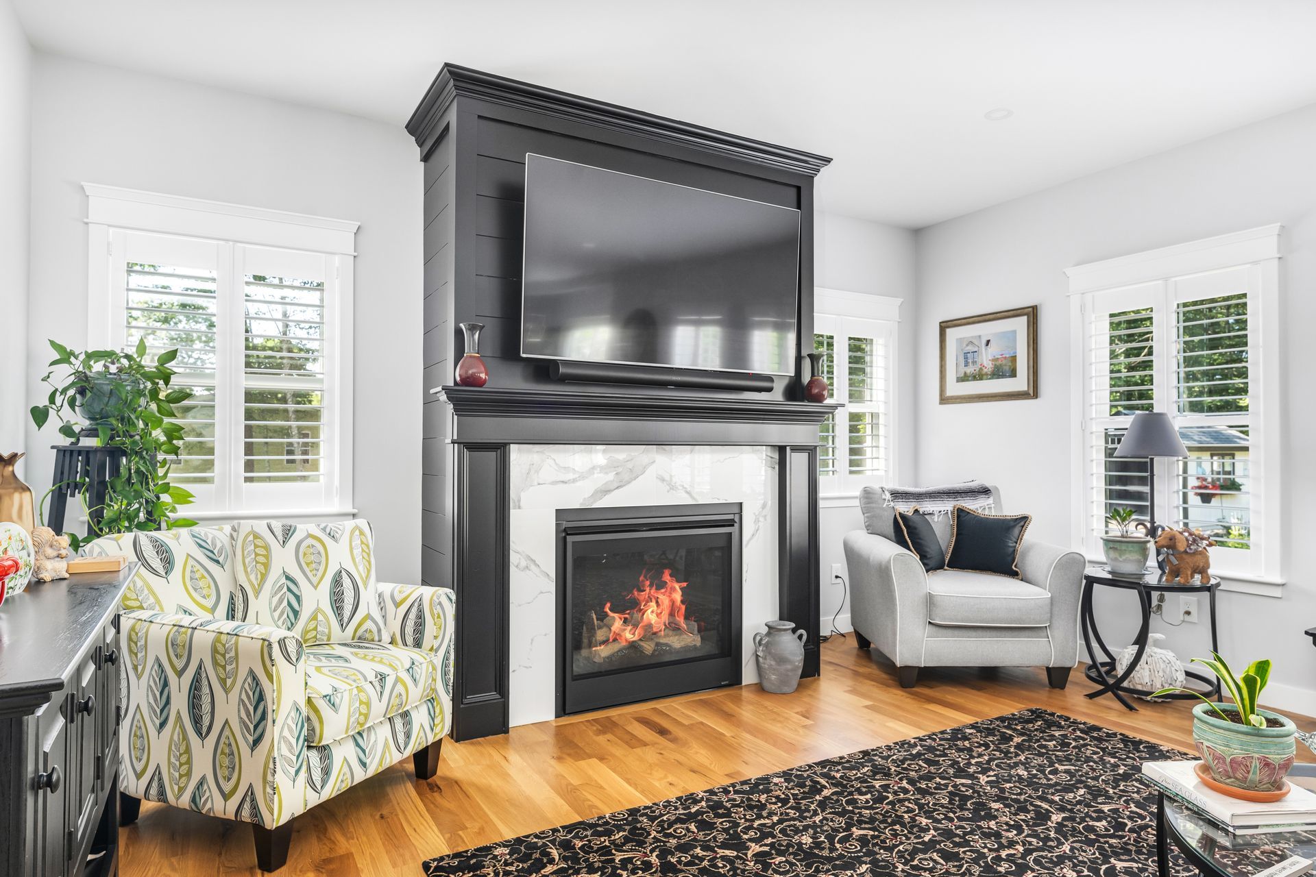 Bright living room with fireplace, black mantel TV, patterned chair, and large windows