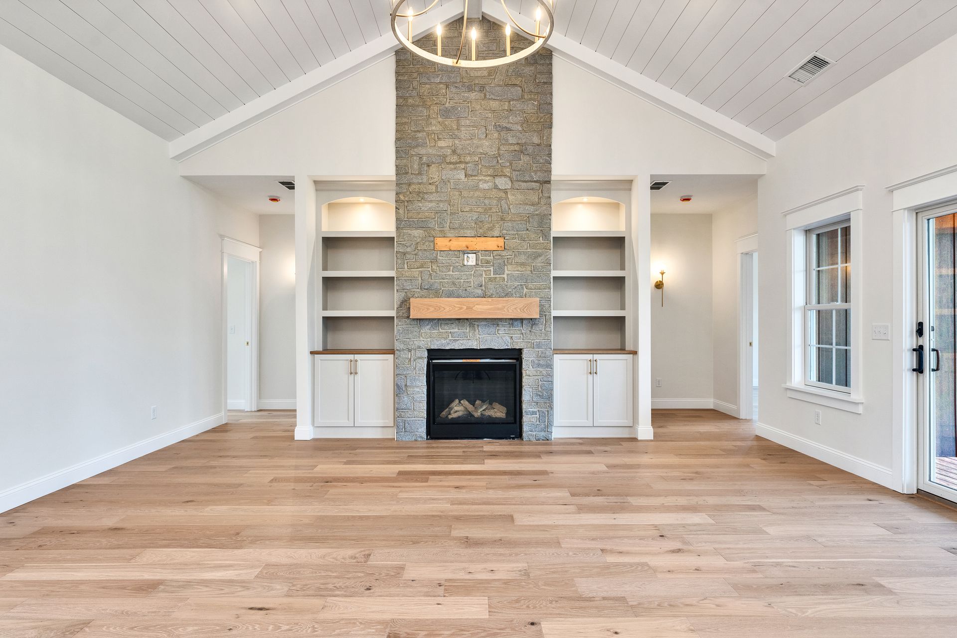 Bright empty living room with vaulted ceiling, stone fireplace, built-in shelves, and hardwood floors