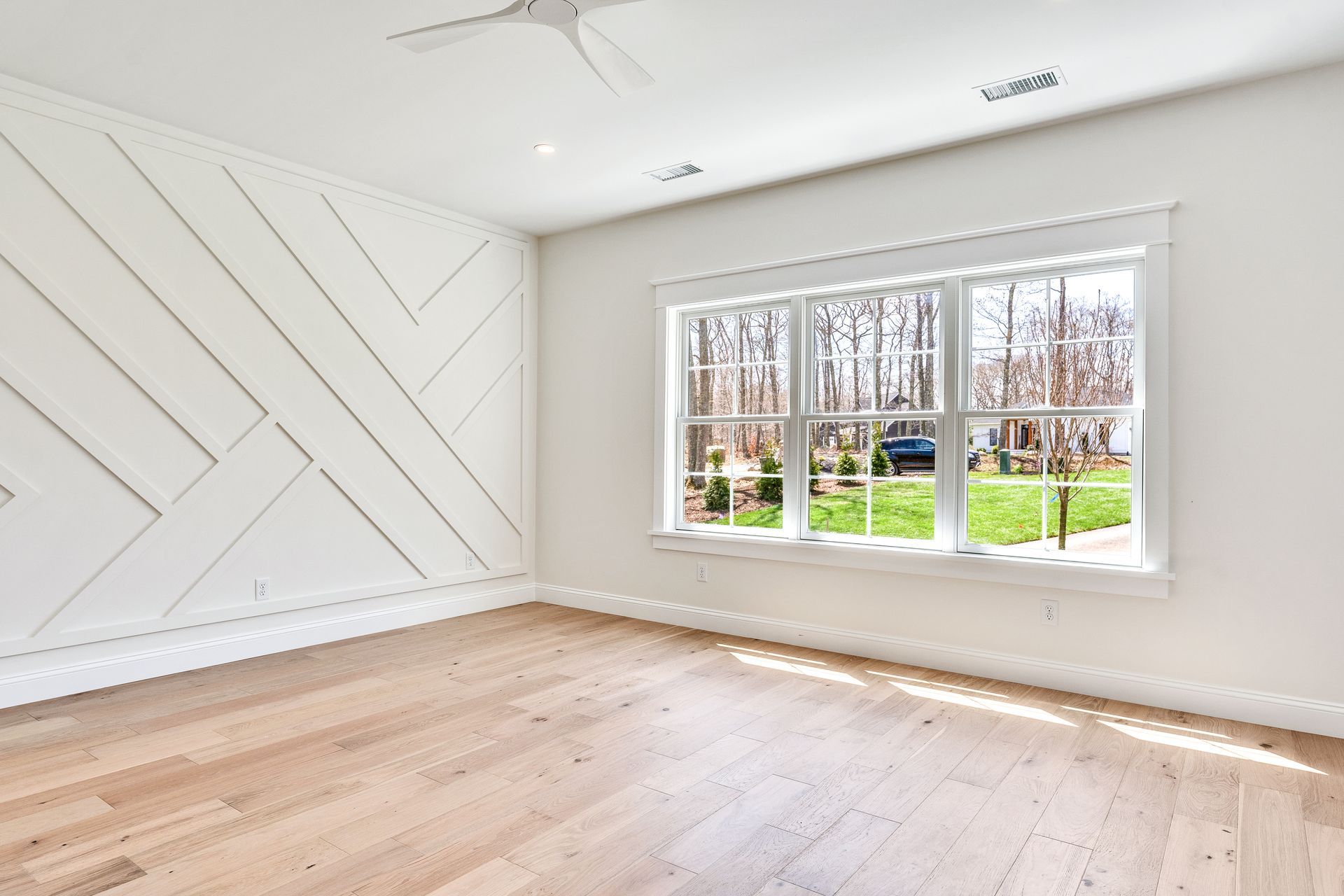 Bright empty room with white walls, light wood floors, and a large window overlooking greenery