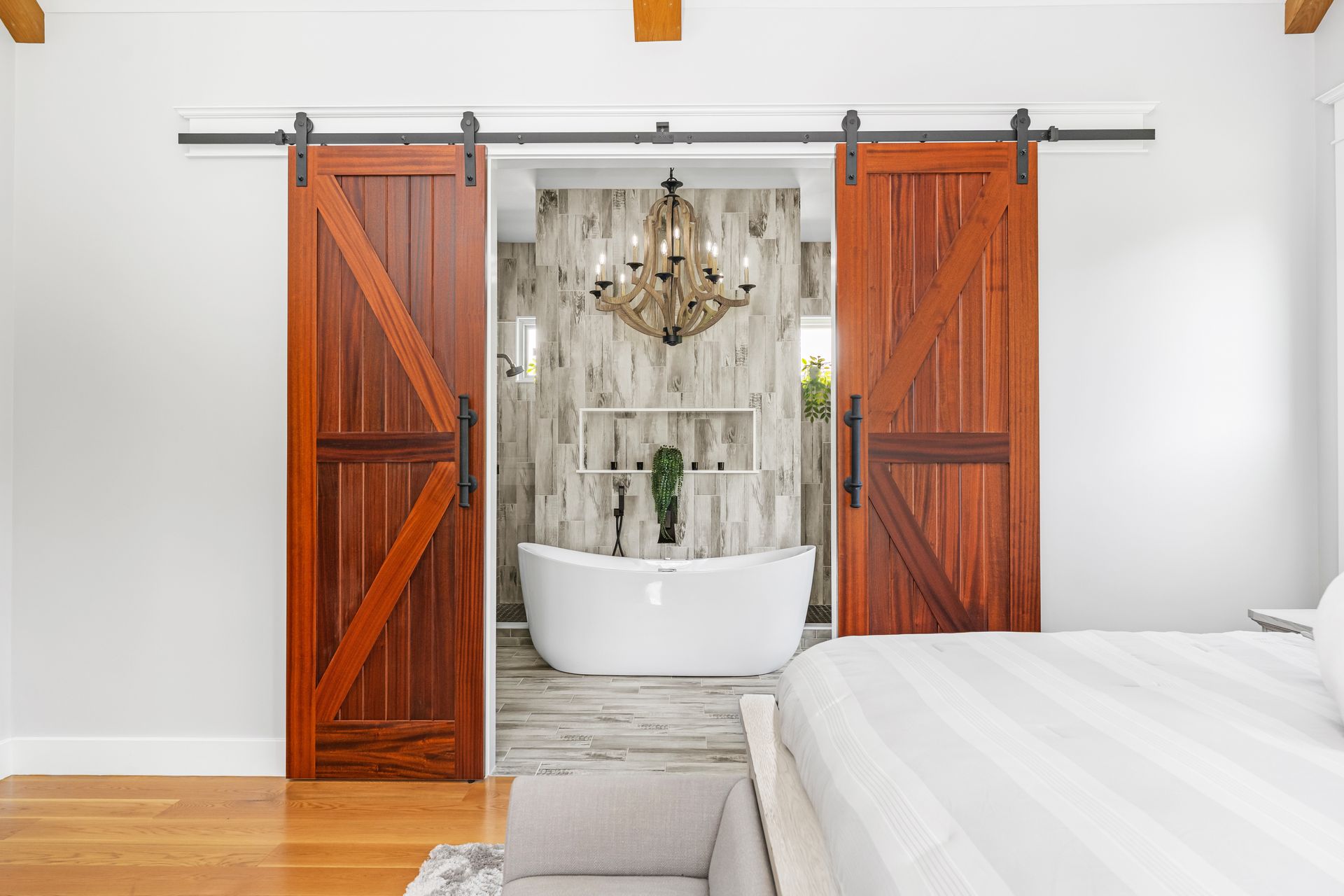 Rustic barn doors open to a freestanding tub beneath a chandelier in a bright, modern bedroom