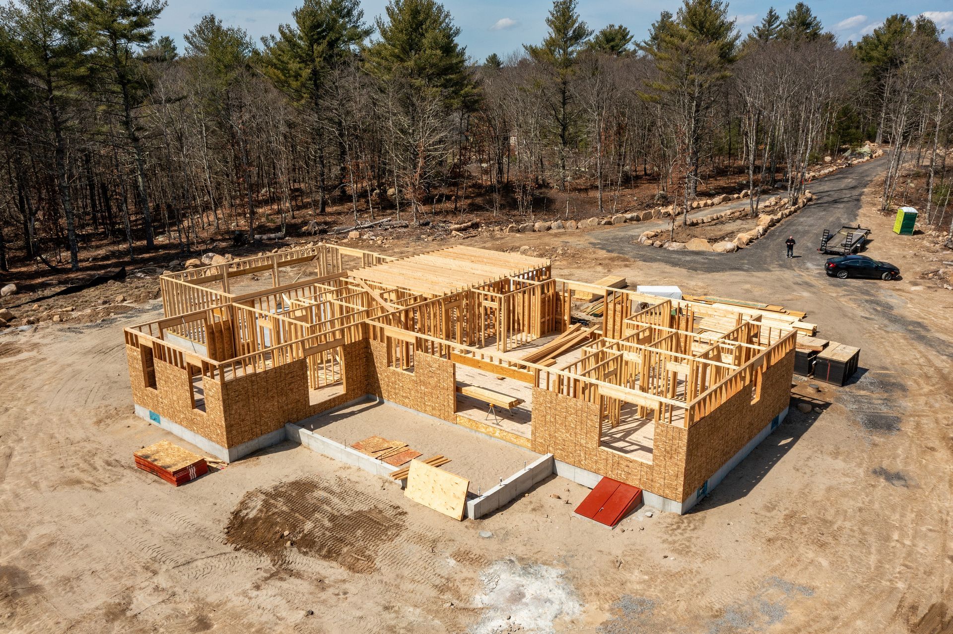 Partially built wooden house framing on a dirt lot surrounded by trees