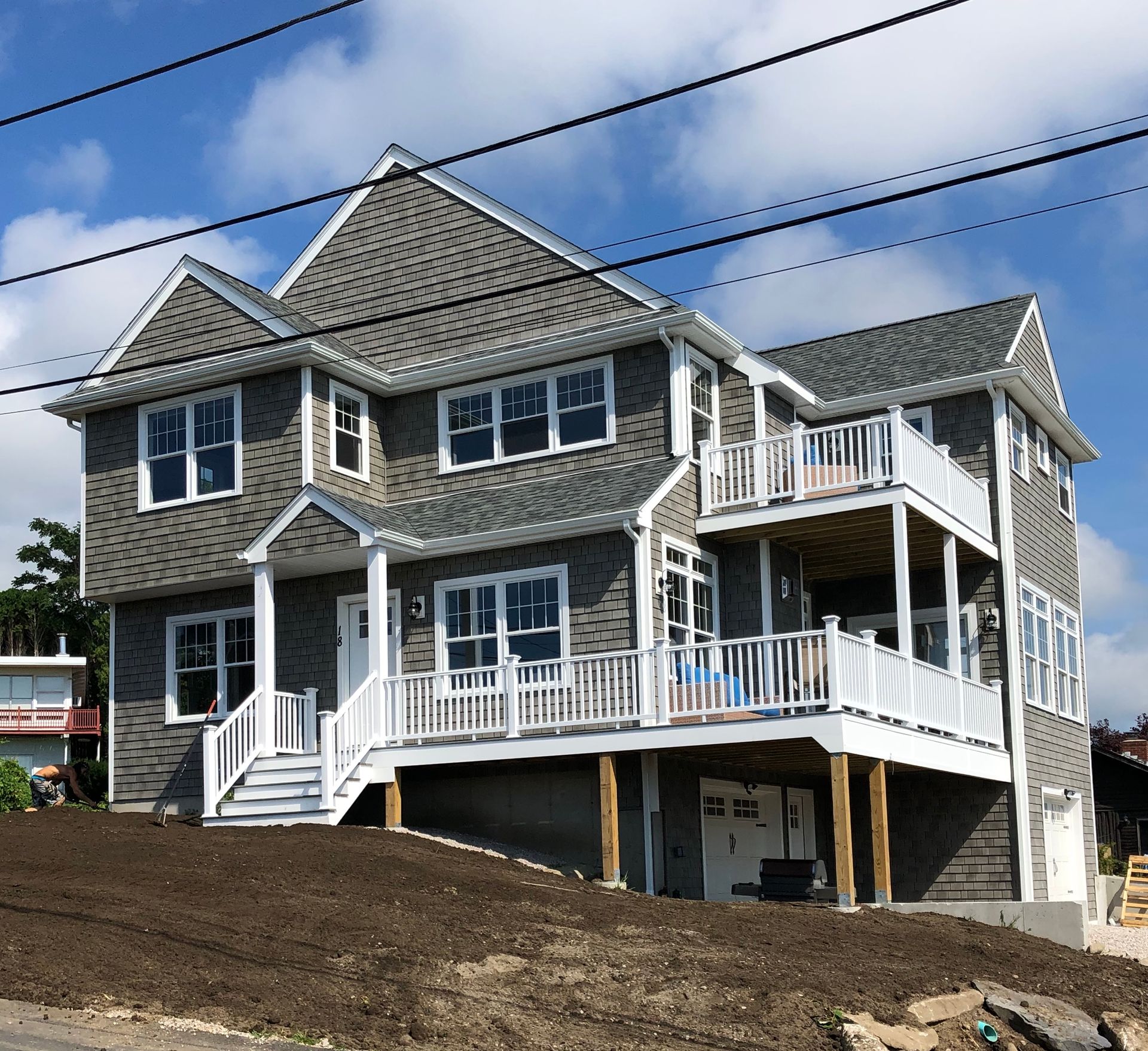 Large gray house with white trim and balconies on a sloped dirt lot under a blue sky