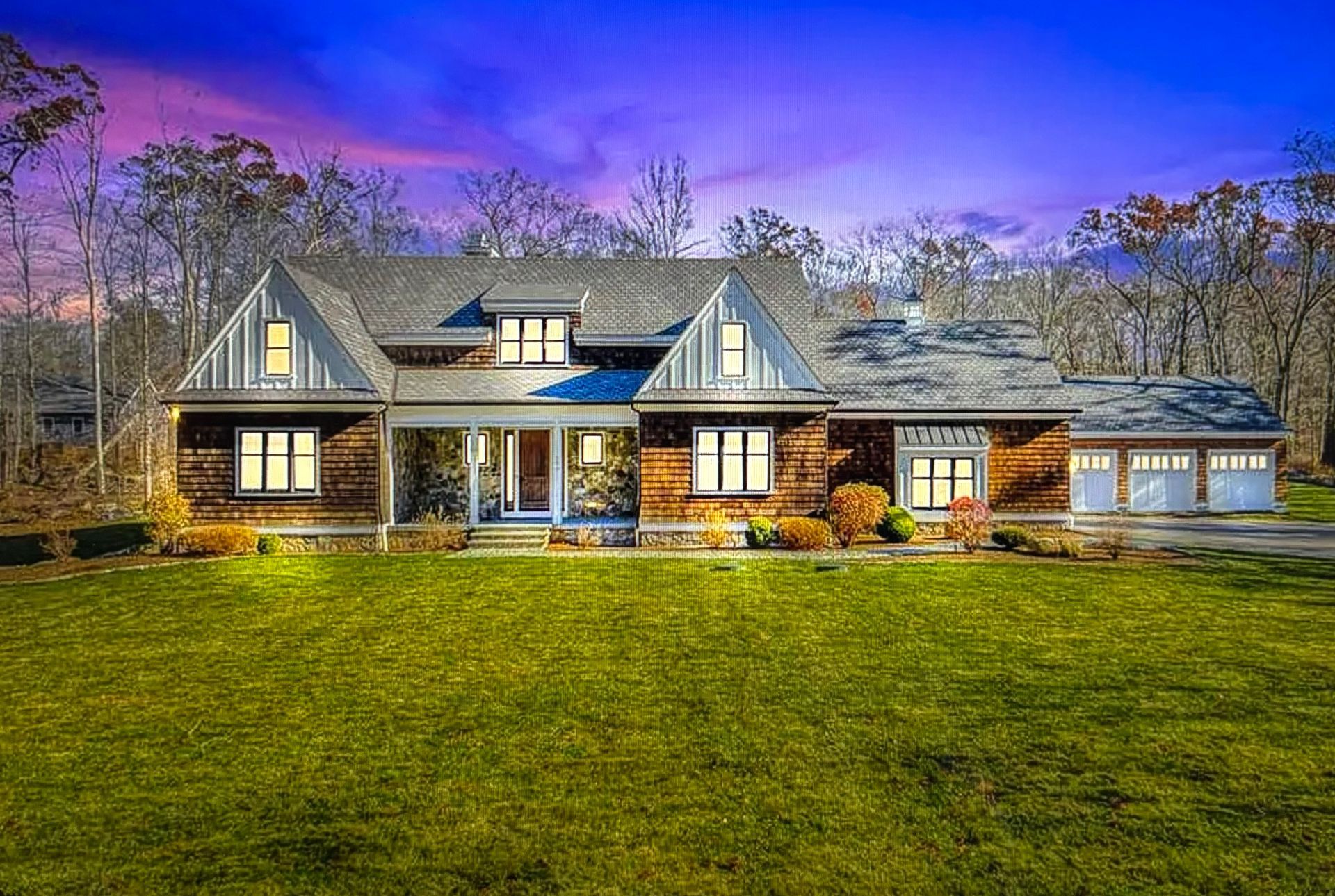 Large stone house with lit windows and a wide green lawn at dusk under a purple sky