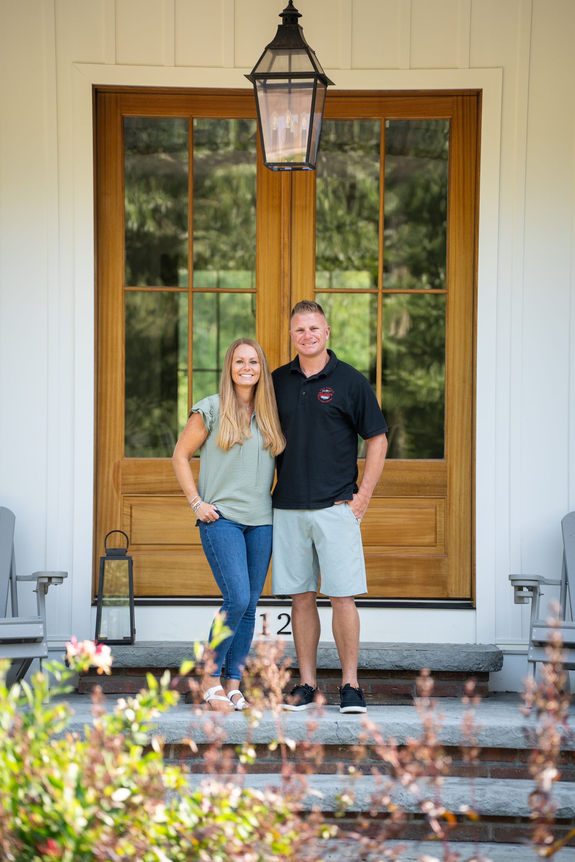 A couple stands on a porch in front of a wooden door with a hanging lantern.