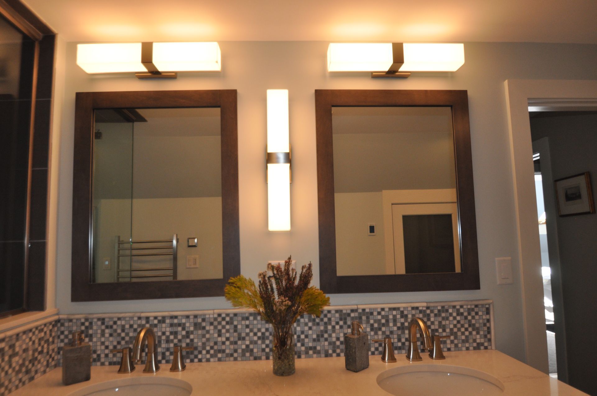 Bathroom with two framed mirrors, sinks, and lighting fixtures. Light blue walls, and a mosaic tile backsplash.