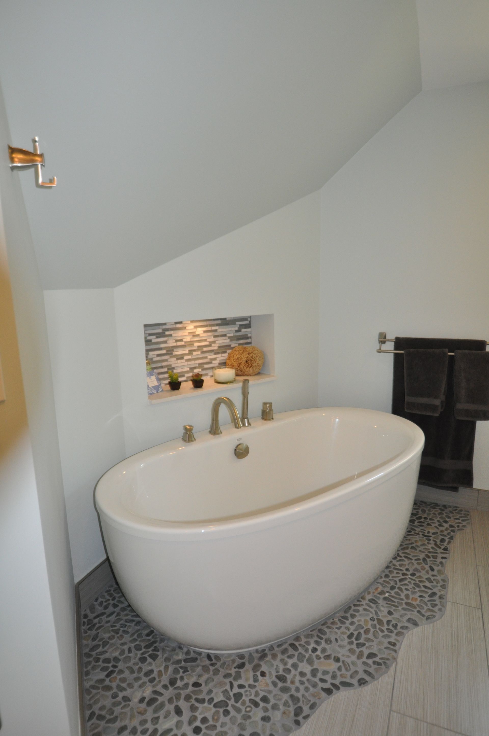 Oval bathtub in a minimalist bathroom with a pebble floor and tiled niche in the slanted ceiling.