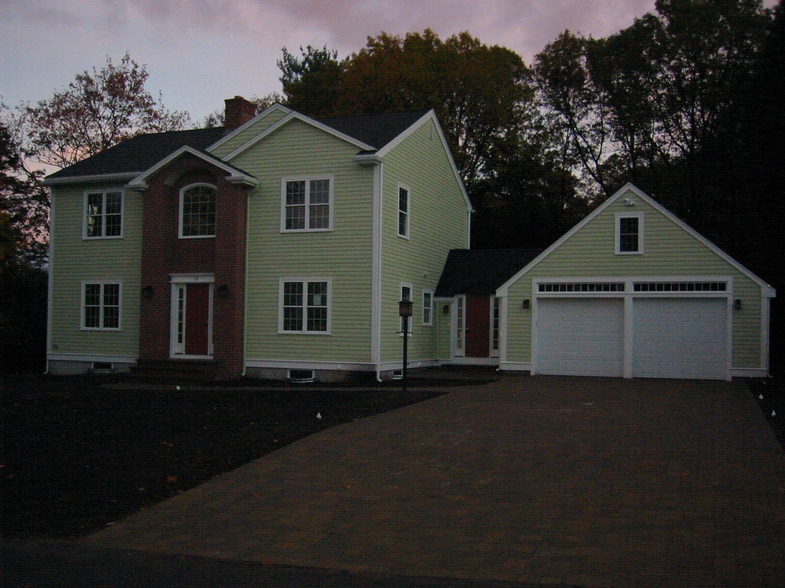 Two-story house with a garage, light green siding, brick-red entrance, and dark driveway at dusk.
