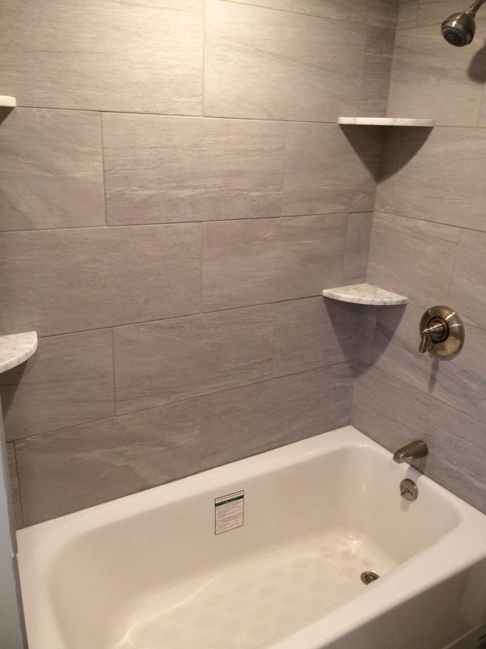 Bathroom with a white bathtub, gray tiled walls, and white shelves.