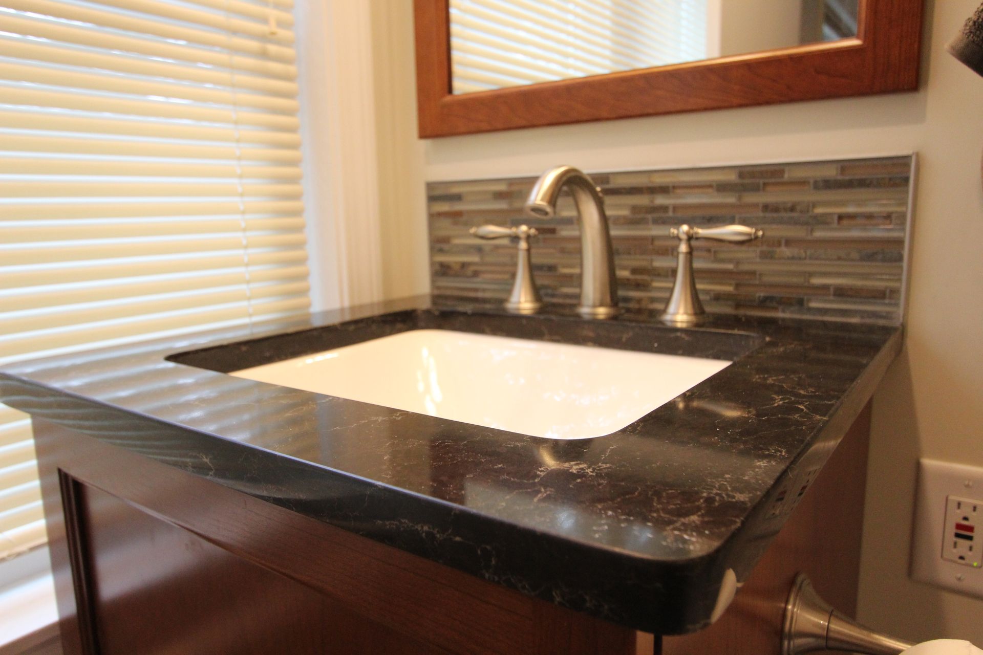 Dark countertop and sink, with tiled backsplash and a wood-framed mirror in a bathroom.