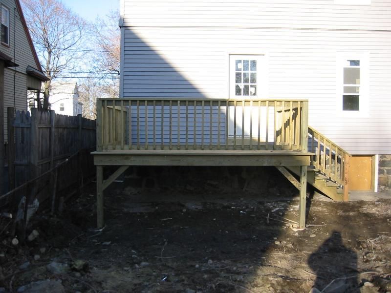 Wooden deck attached to a light-colored house with a railing, supported by wooden beams, in a backyard.