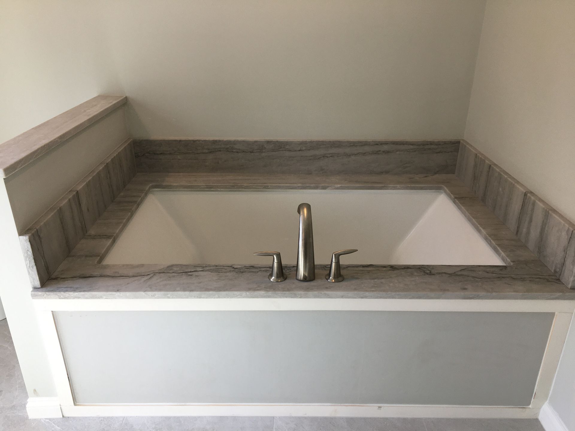 Built-in bathtub with marble countertop and silver faucet, set in a bathroom with gray and white walls.