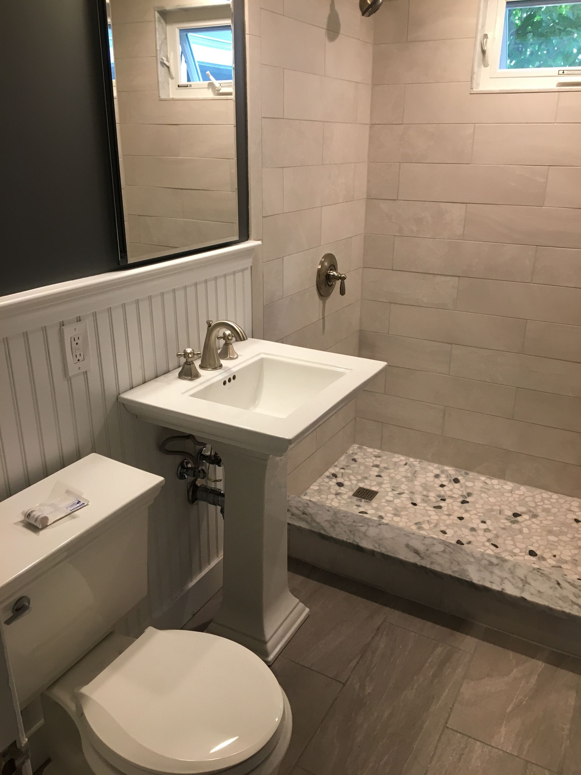 Bathroom with a pedestal sink, toilet, and shower with bench. White, gray, and beige tones.