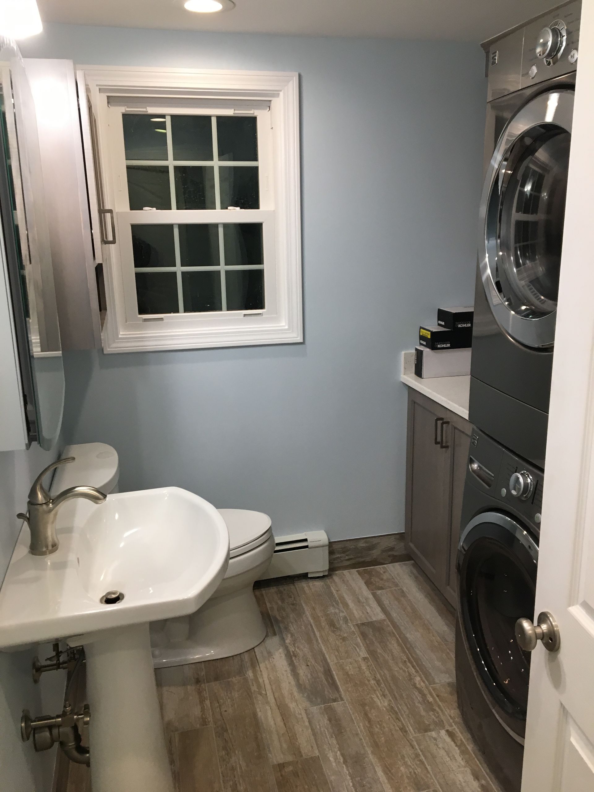 A small bathroom with a pedestal sink, toilet, stacked washer and dryer, and a window. Light blue walls, wood-look flooring.