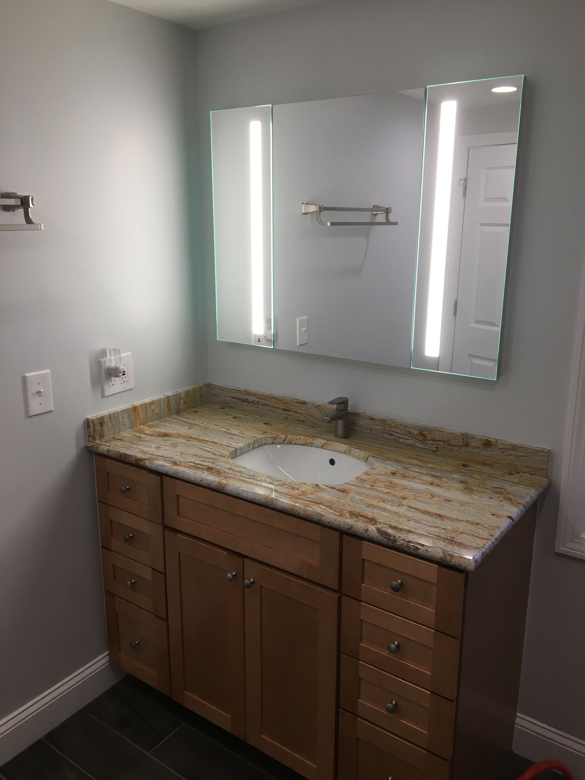 Bathroom vanity with a light wood cabinet, granite countertop, and mirror with lights.