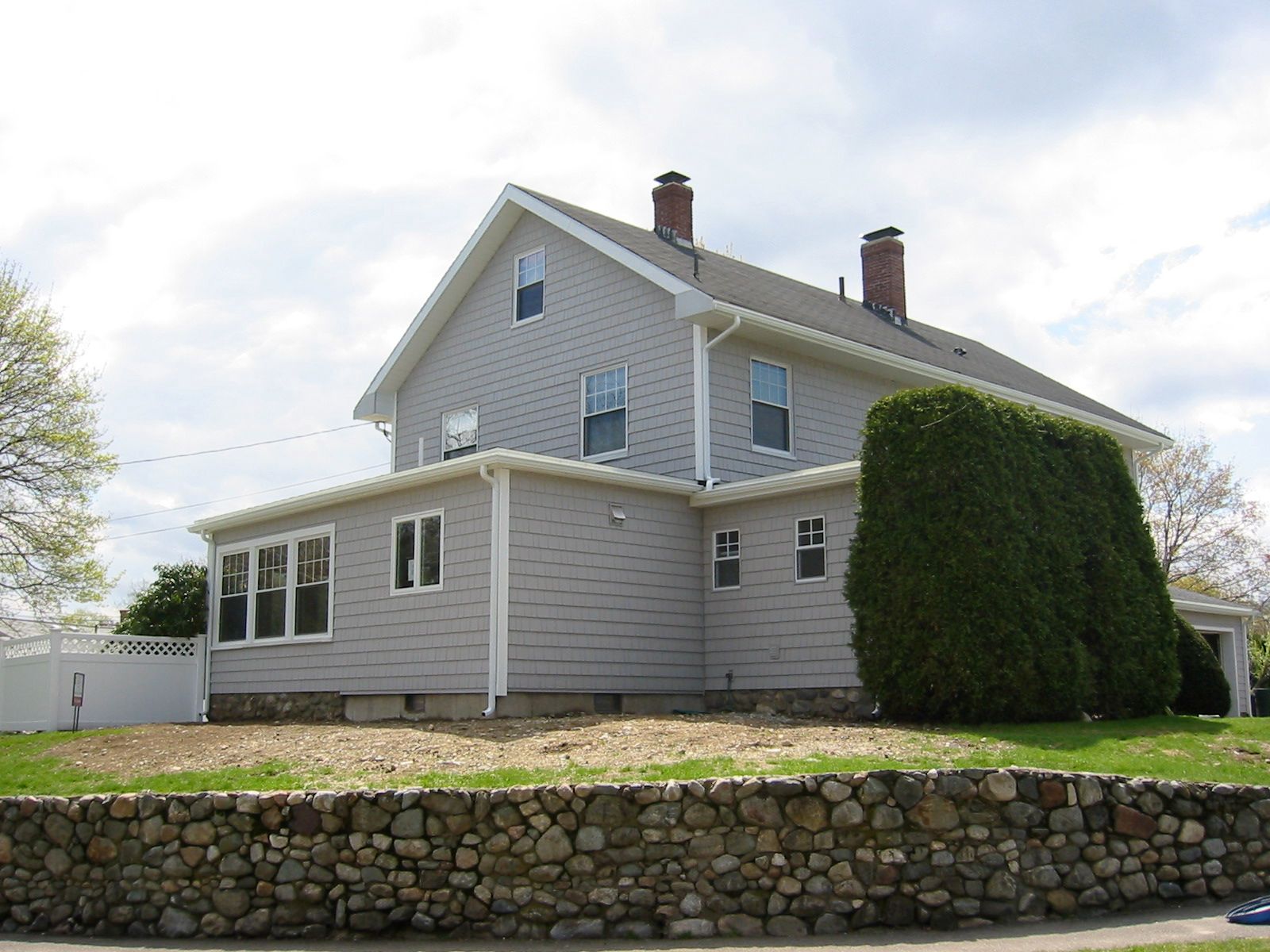 Two-story gray house with a stone wall in front. Overcast sky.