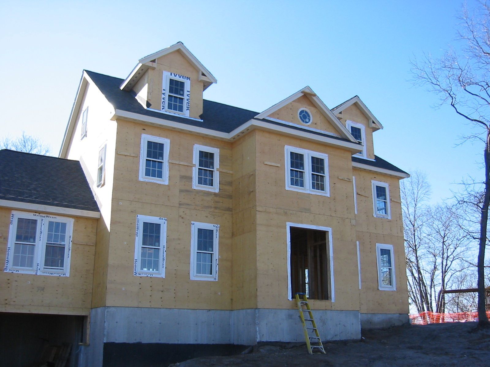 Two-story house under construction with exposed wood siding and multiple windows. Blue sky overhead.