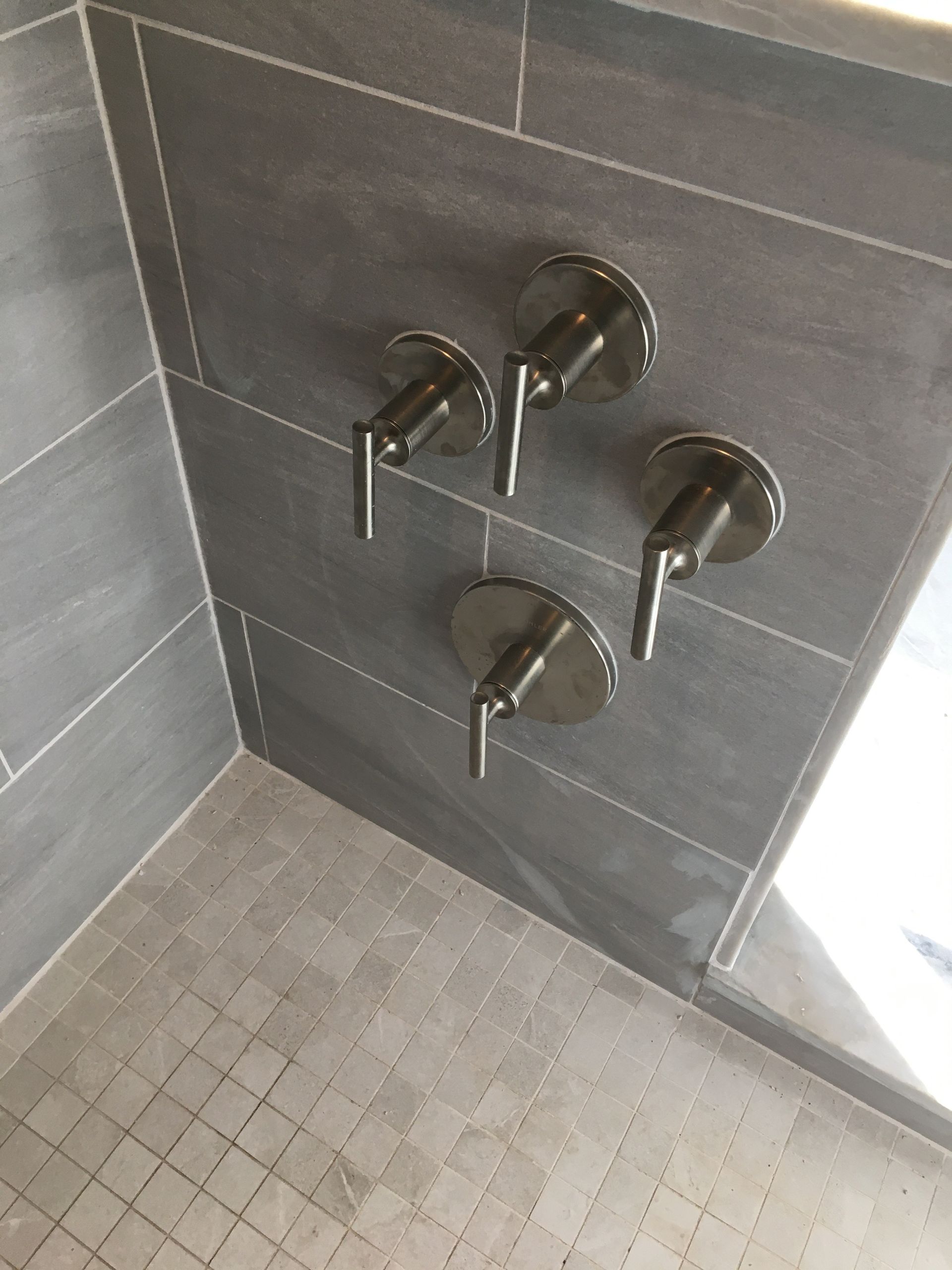 Four silver shower control handles on gray tiled wall.