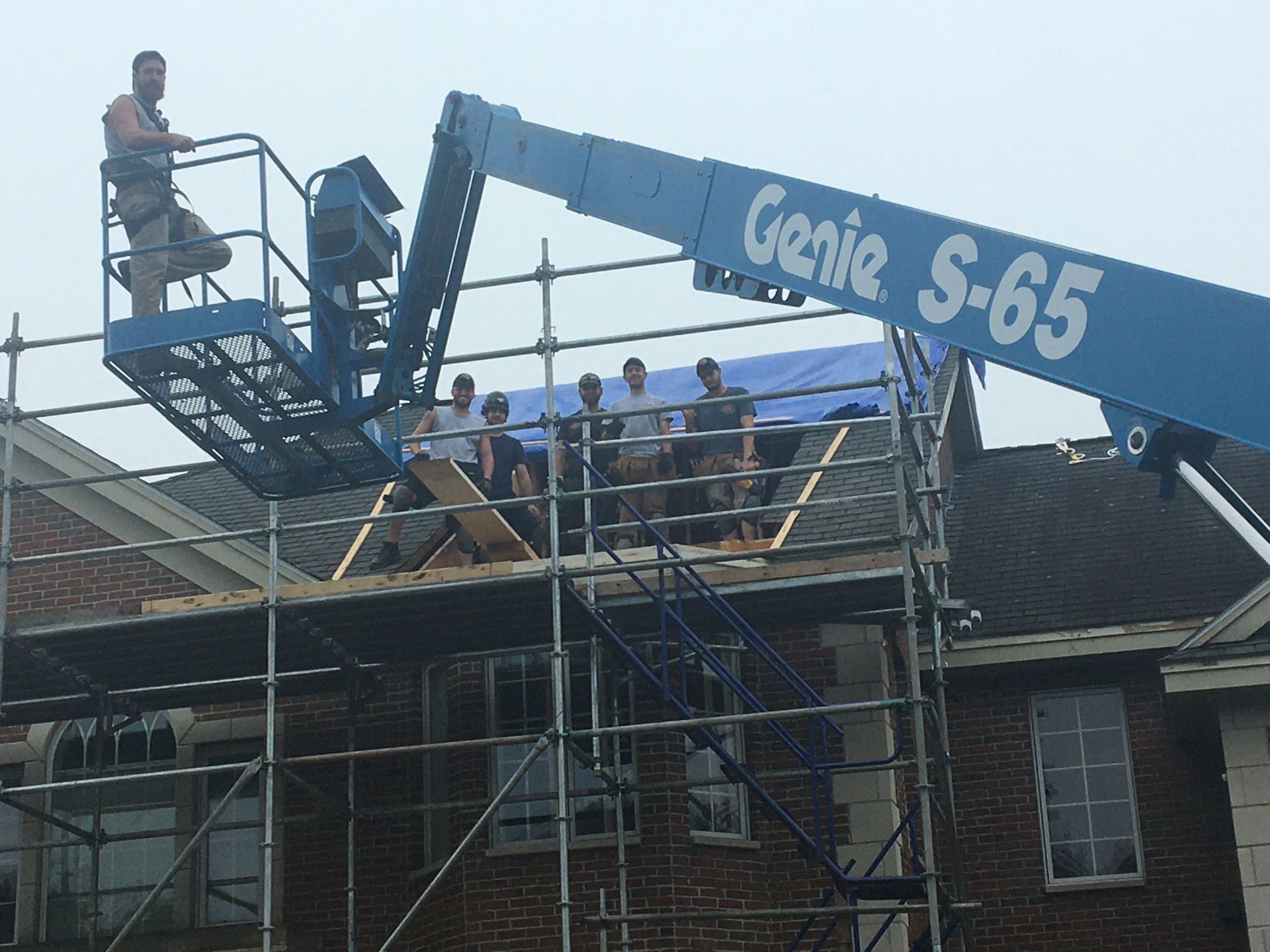 Construction workers on scaffolding, using a lift to work on a roof. Blue lift, brick building, overcast sky.