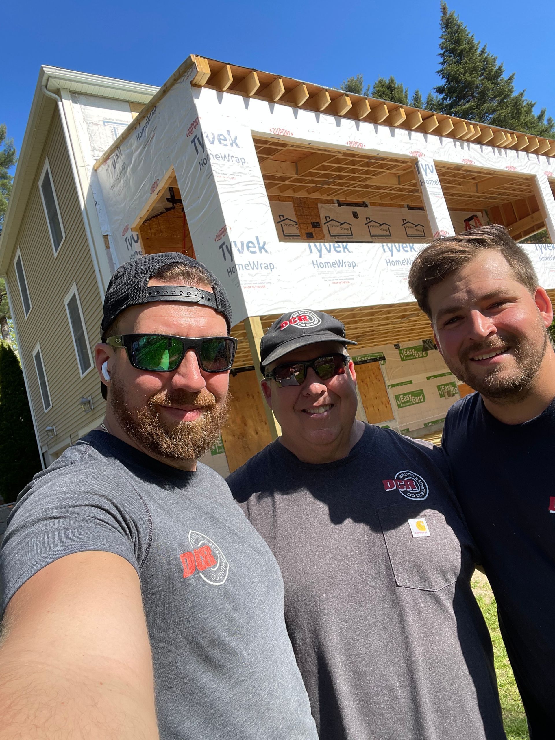 Three men posing for a selfie in front of a house under construction; sunny day.