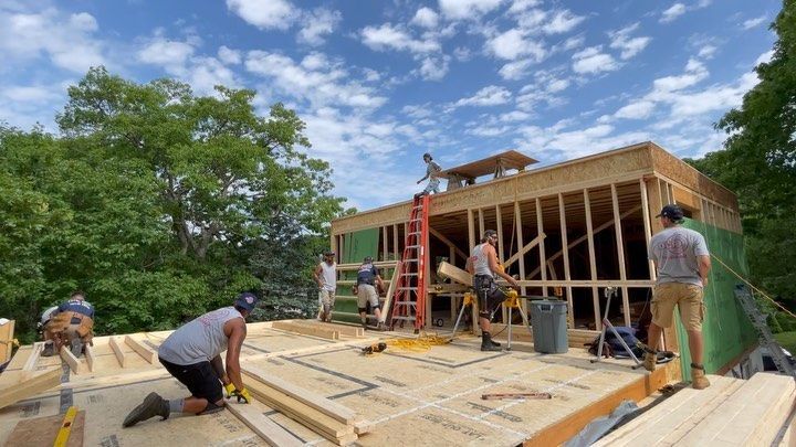 Construction workers building a wood-framed structure outdoors under a partly cloudy sky.