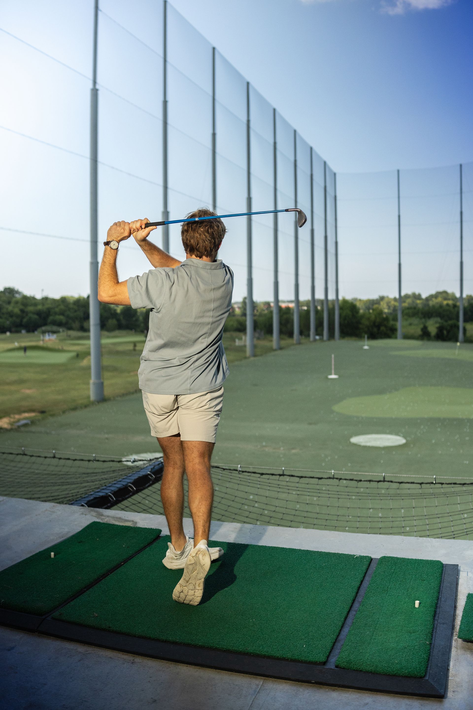 Golfer practicing a swing at a driving range, seen from behind on a green mat.