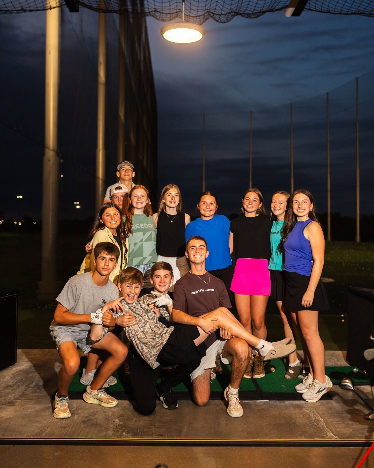Group of people posing on a baseball dugout at dusk, with a bright stadium light overhead.