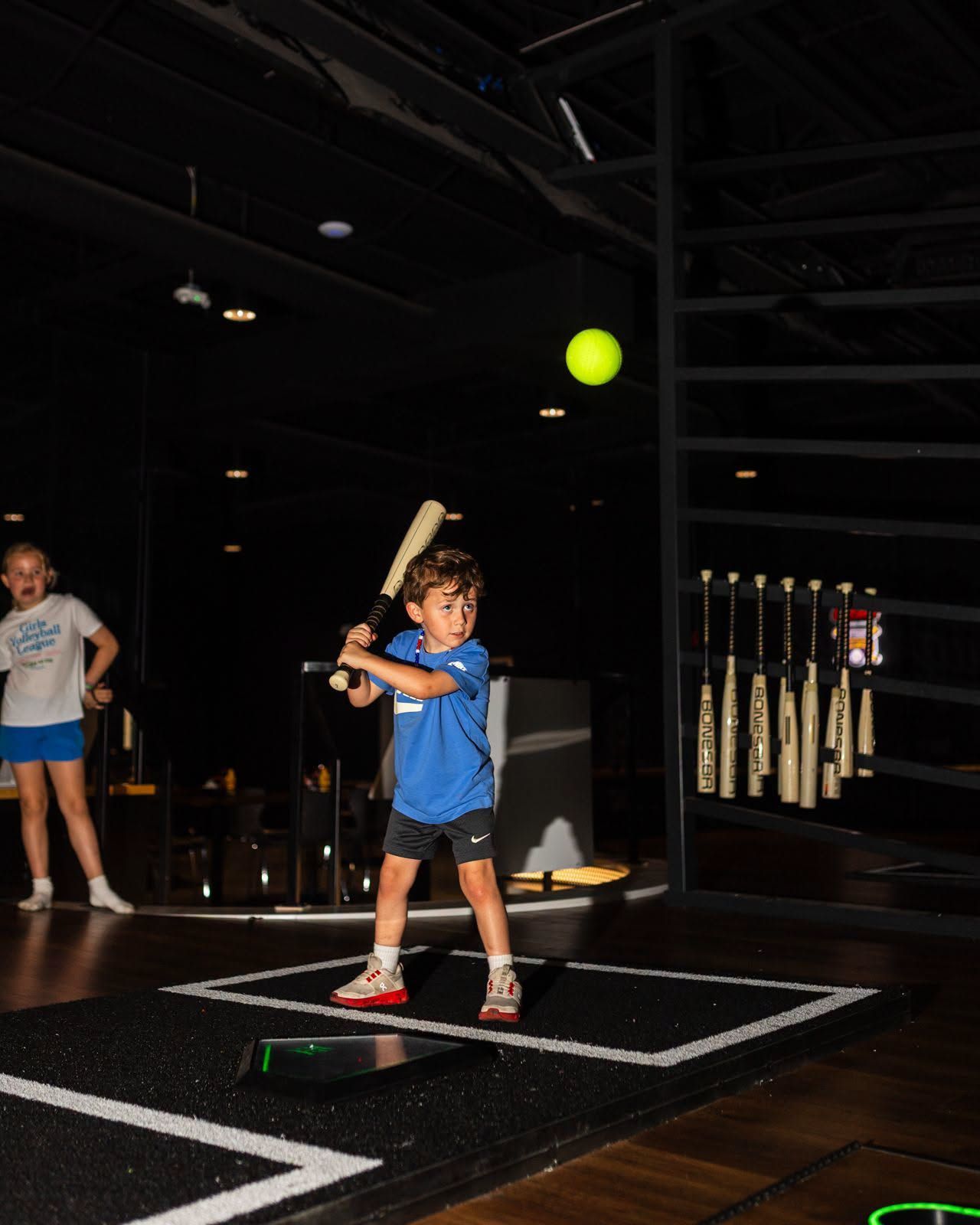 Child swinging a bat at a green ball in an indoor batting cage
