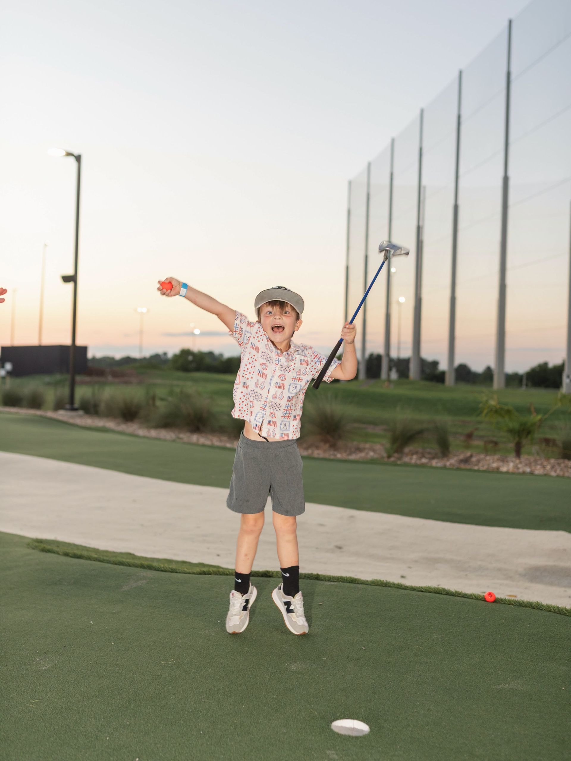 Child jumping on a golf putting green at sunset, holding a club with arms raised.