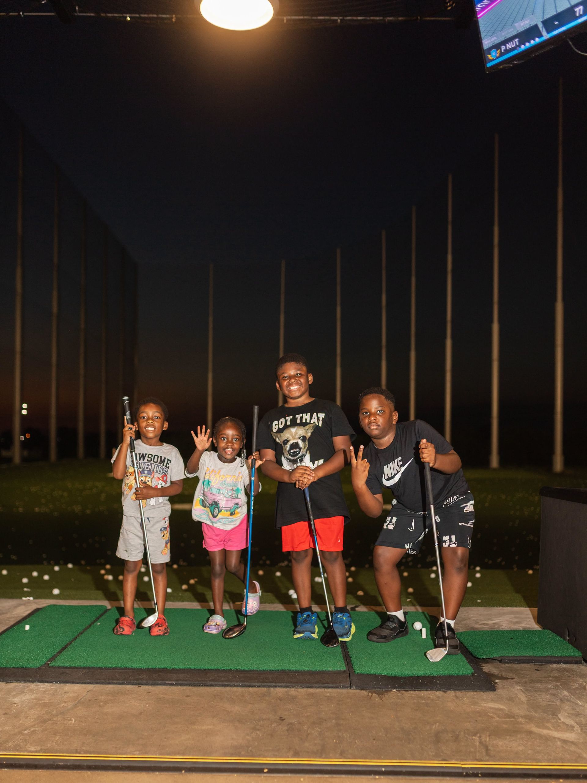 Four children pose at a driving range under a bright overhead light, with golf nets in the background.