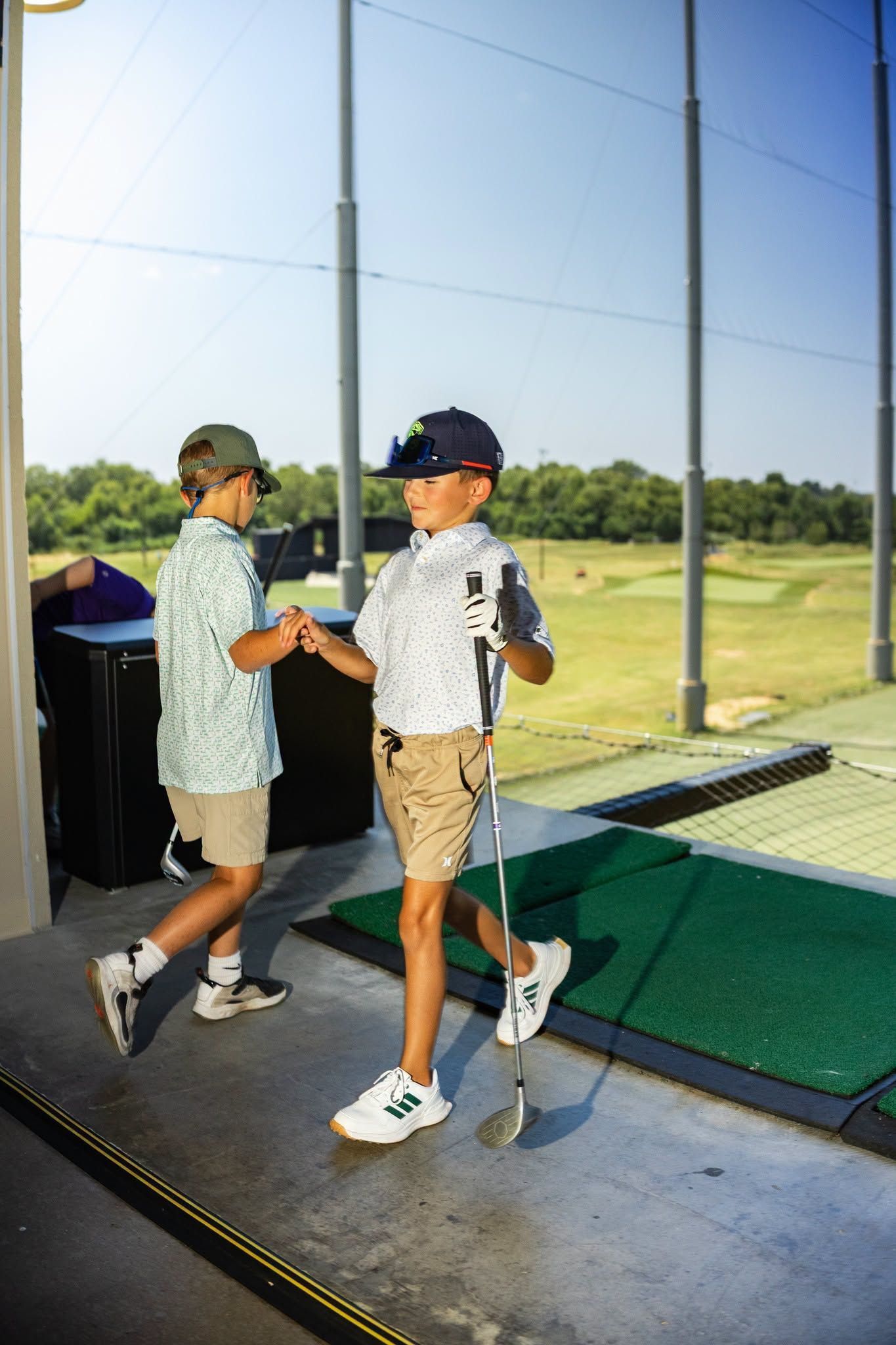 Two children in golf attire stand at a driving range beside green hitting mats.