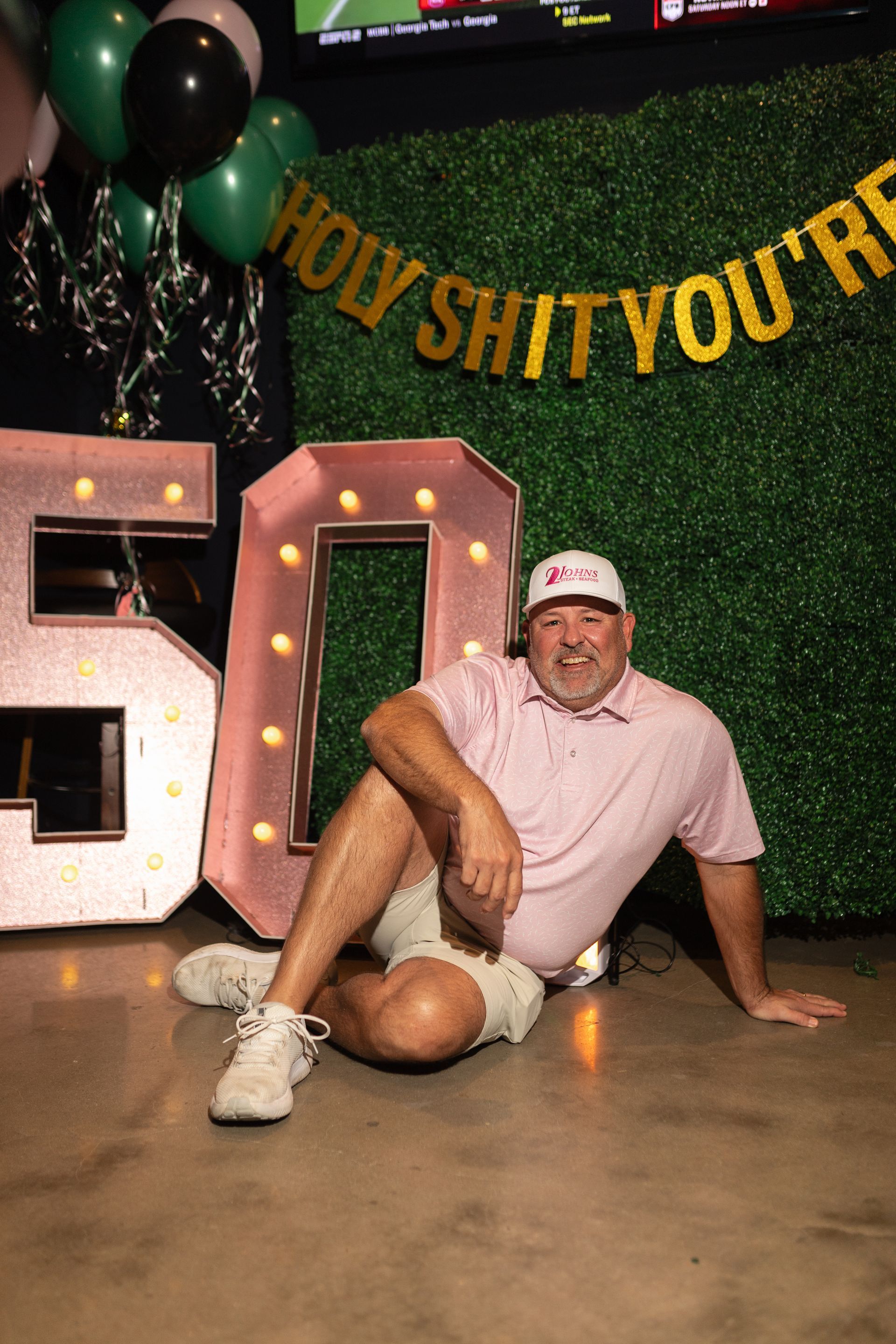 Man seated near a "50" sign, green backdrop, balloons and "Holy Shit You're" banner.
