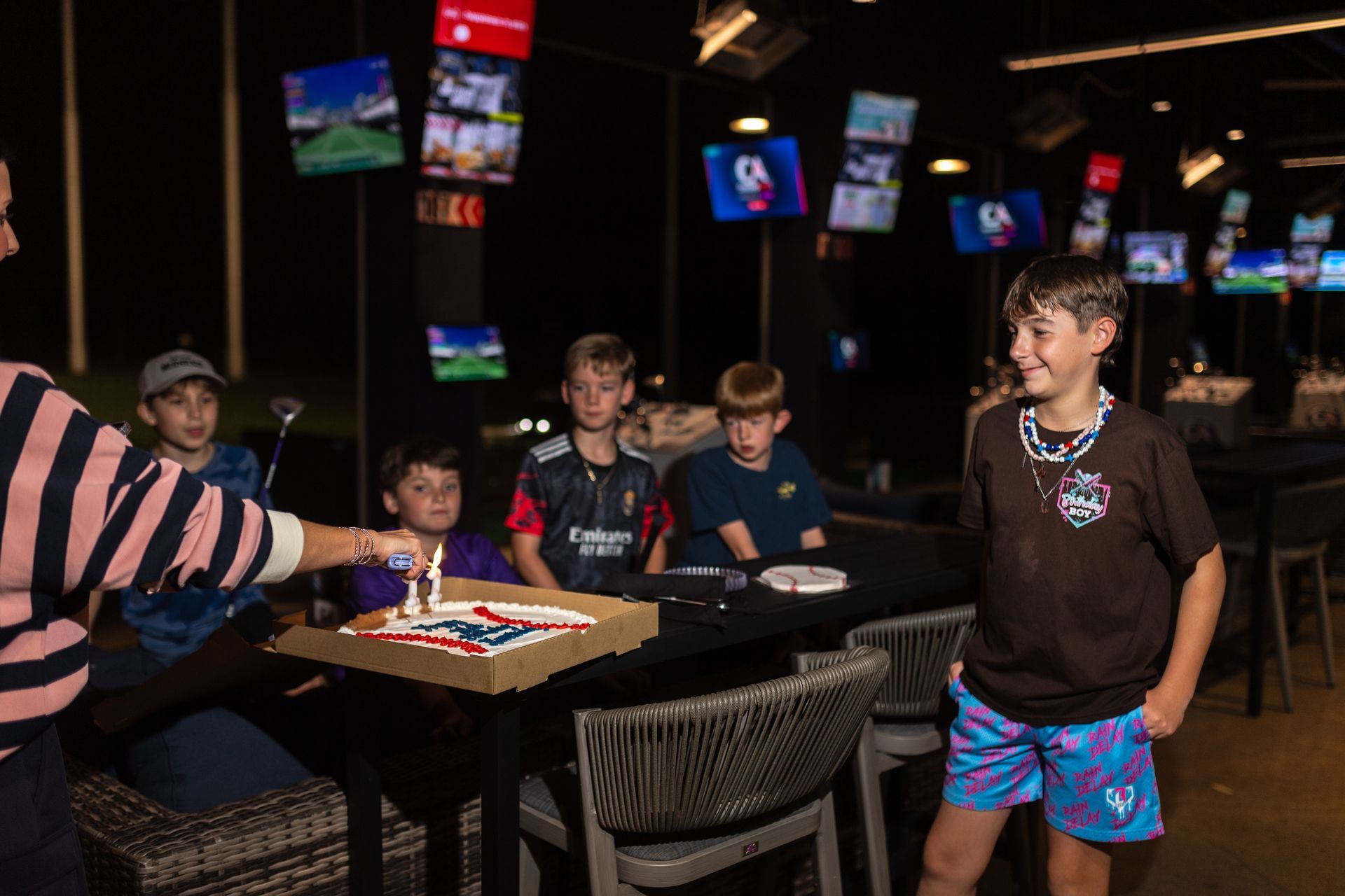 Group of children celebrating with a decorated cake at a venue with screens.