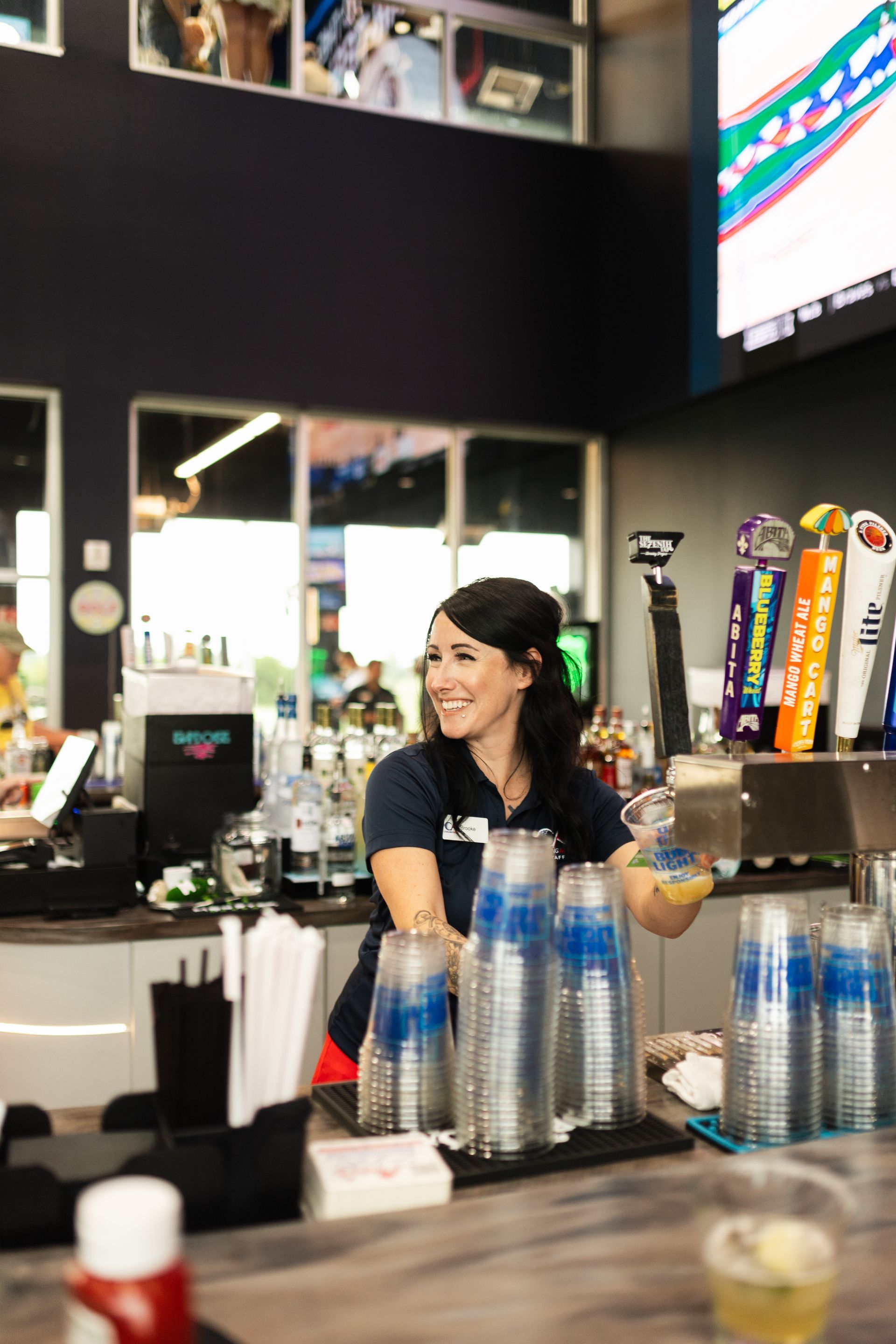 Bartender smiling behind a bar, with bottles and stacked cups in front of her.