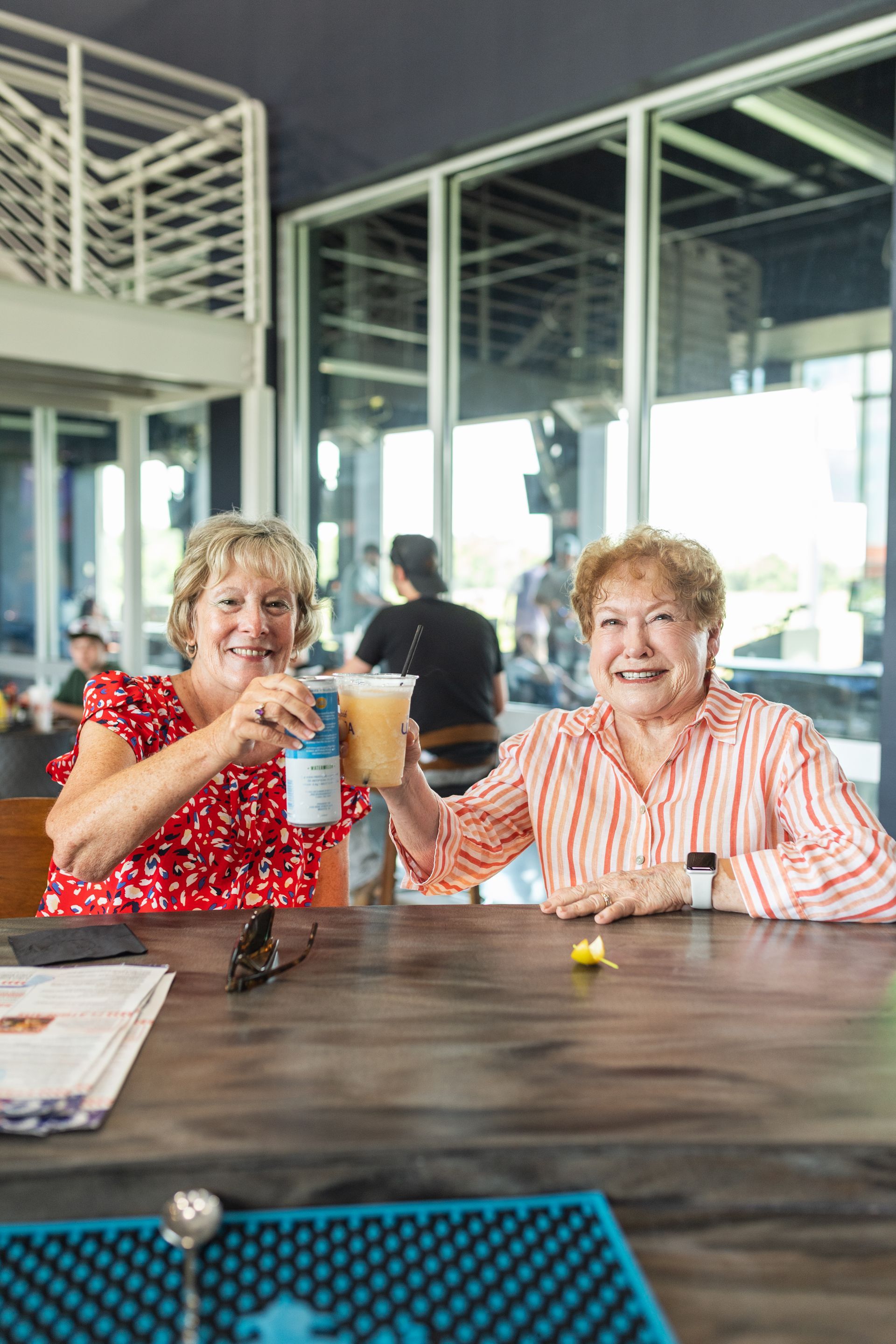 Two people smiling and toasting drinks at a café table by large windows