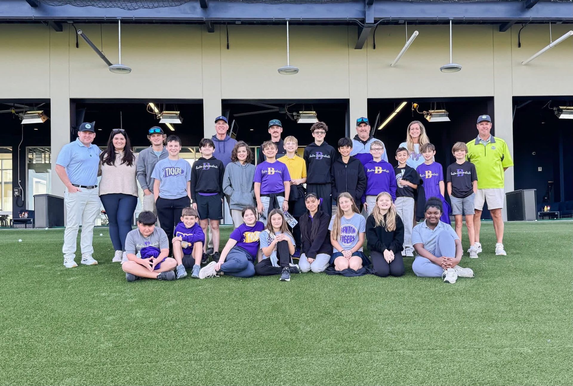A group of people pose for a photo on a green field in front of open garage bays at an outdoor facility.