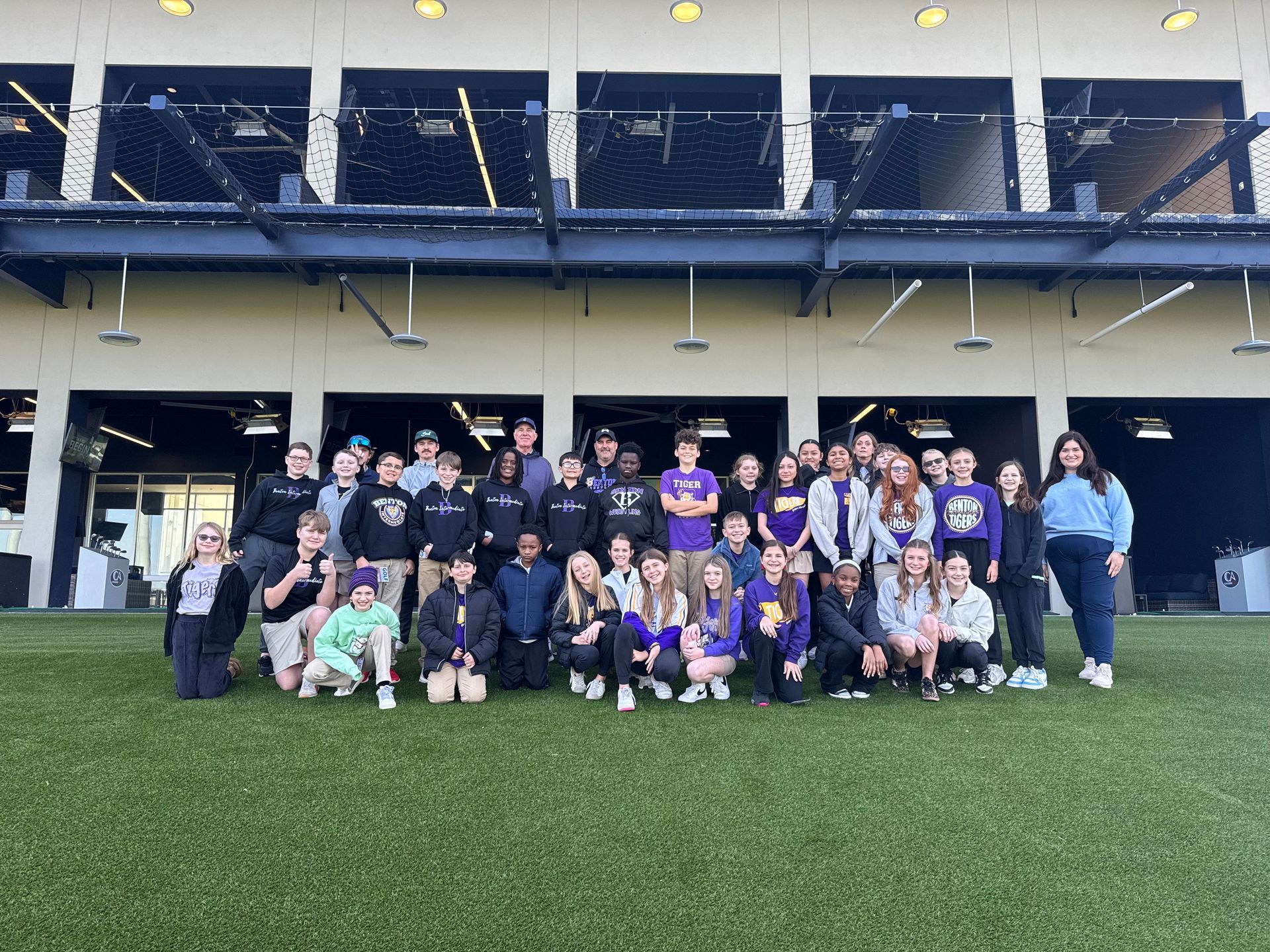 A large group stands in rows on artificial turf in front of a multi-story building at an outdoor sports complex.
