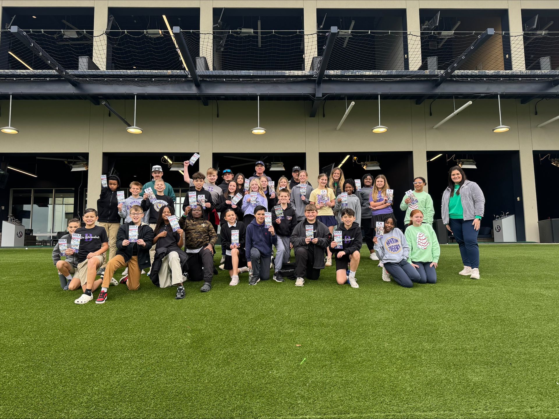 A group poses for a photo on green turf at an outdoor driving range, with many people holding papers.