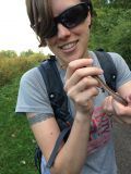 Person smiling, holding a worm outdoors. Wearing sunglasses, backpack, and a grey shirt.