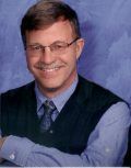 Man in glasses, smiling, wearing a blue shirt, tie, and dark vest; arms crossed against a blue background.