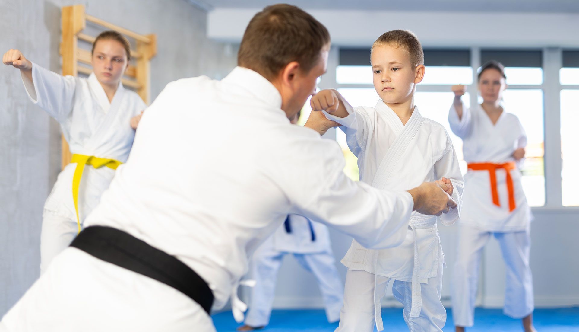 Martial arts class: Instructor guides a student's punch, others practice in the background. White gis, colorful belts.