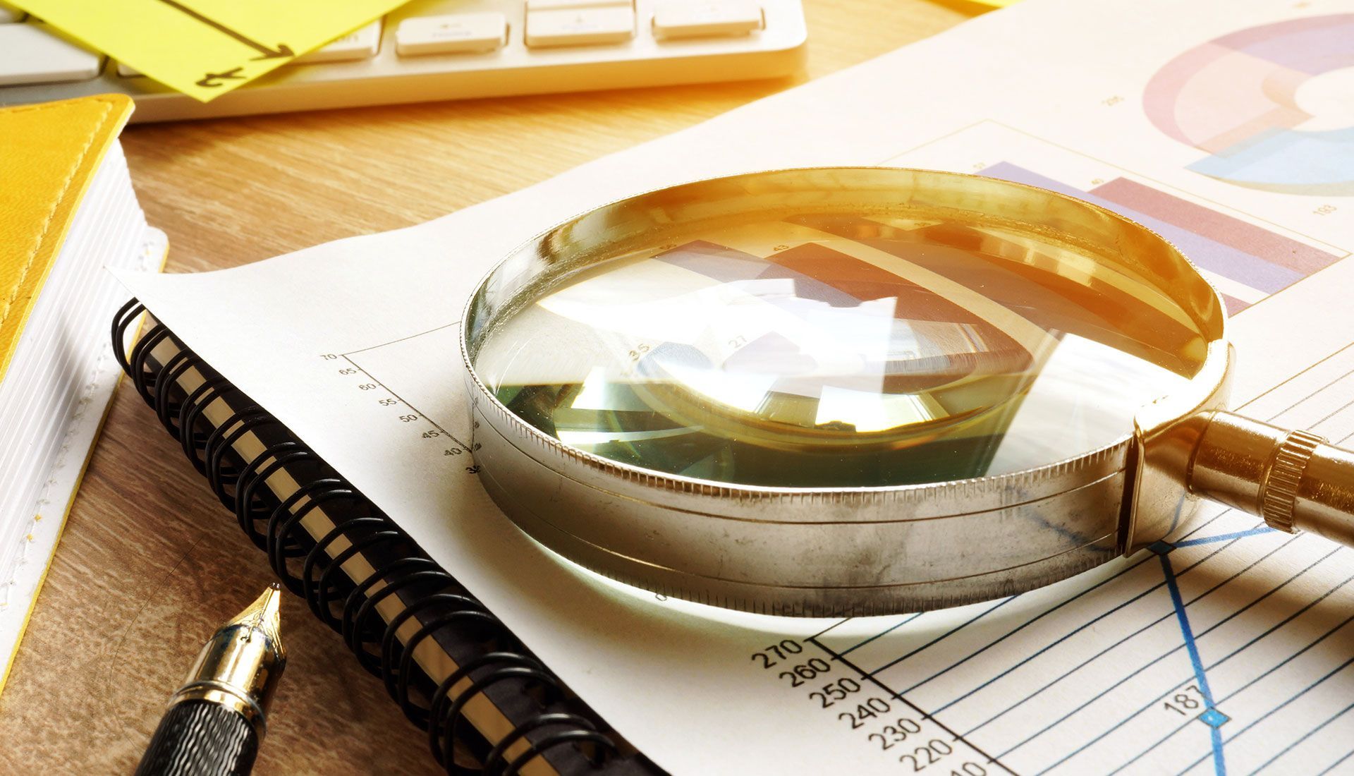 Magnifying glass on financial documents, notebook, pen, and keyboard on a desk.