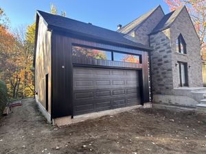 Extension moderne de garage noir attenante à une maison en pierre, avec des arbres d'automne en arrière-plan.